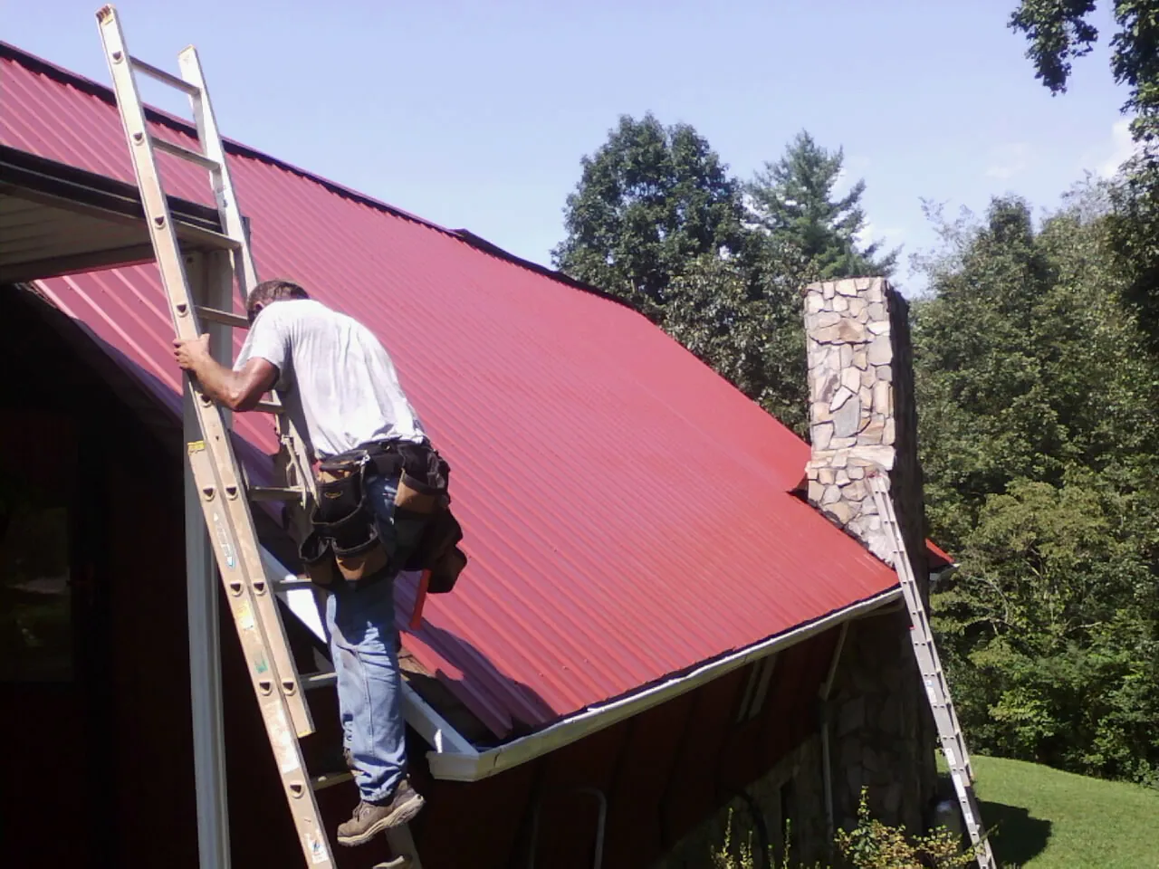 Roofer on a ladder beside a red metal roof and stone chimney — Soffit & Fascia Repair by Roofing Brothers in Mabelvale
